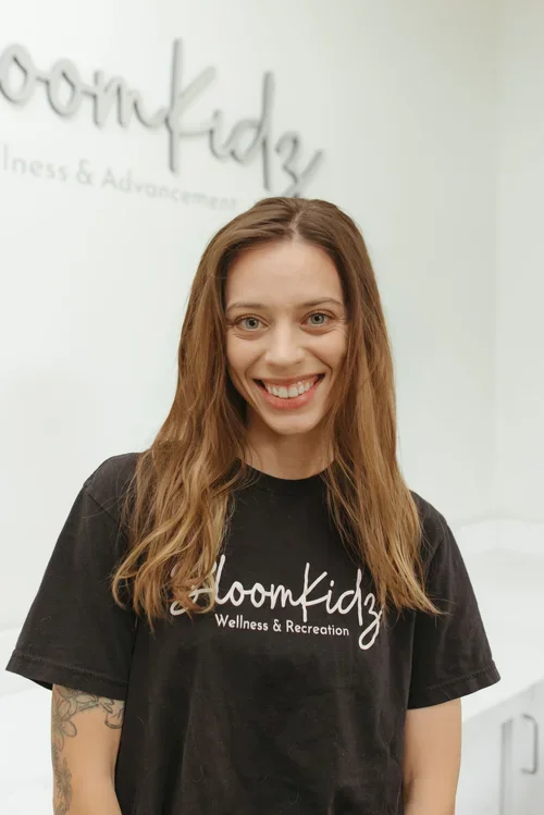 A smiling young woman with long brown hair, wearing a black t-shirt with 'BloomKidz Wellness & Recreation' printed on it, standing in front of a white wall with the same logo in the background.