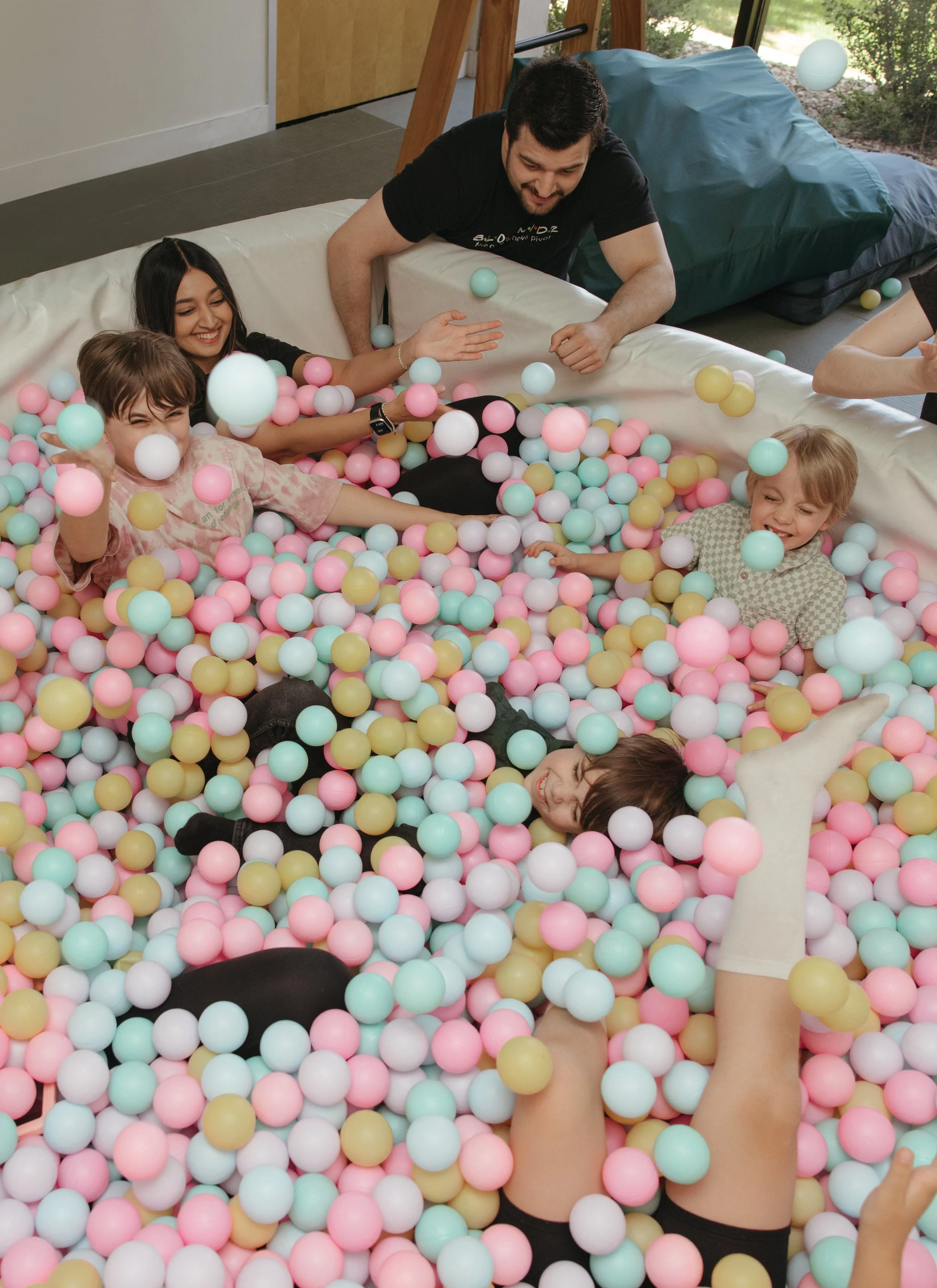 People playing in a colorful ball pit with pink, yellow, white, and mint green balls, with a window and scenery outside in the background.