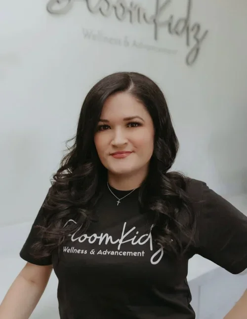 Woman with long dark hair smiling at camera, wearing a black t-shirt with 'BloomKidz Wellness & Advancement' written on it, standing in front of a wall with similar text.
