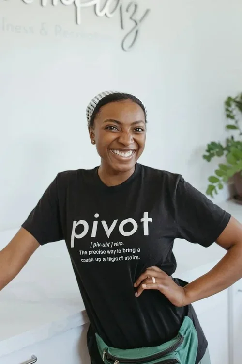 A smiling woman wearing a black T-shirt with the word "pivot" and a definition, standing indoors near a white counter with a plant in the background.