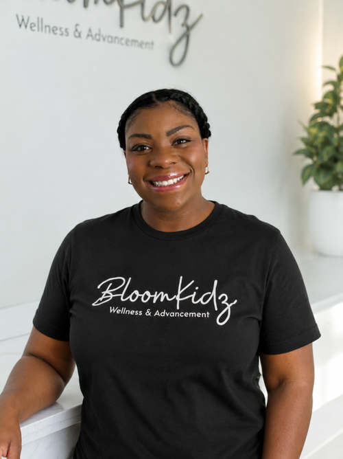 A smiling woman wearing a black t-shirt with 'Bloomkidz Wellness & Advancement' printed on it, standing in a wellness center with a white wall and a potted plant in the background.