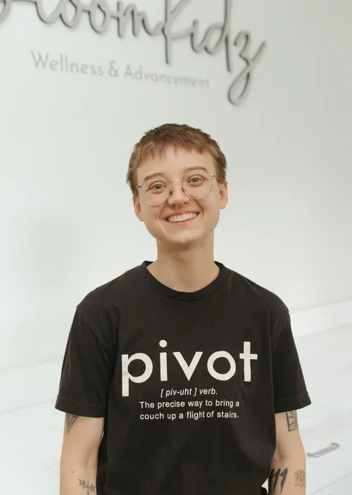 Smiling person wearing glasses and a black t-shirt with the word "pivot" on it, standing in front of a white wall with a sign that says "Wellness & Advancement."