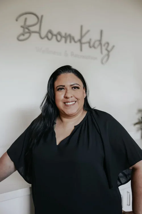 Woman with dark hair and a black top smiling in front of a blurred sign that reads 'BloomKidz Wellness & Advancement'.