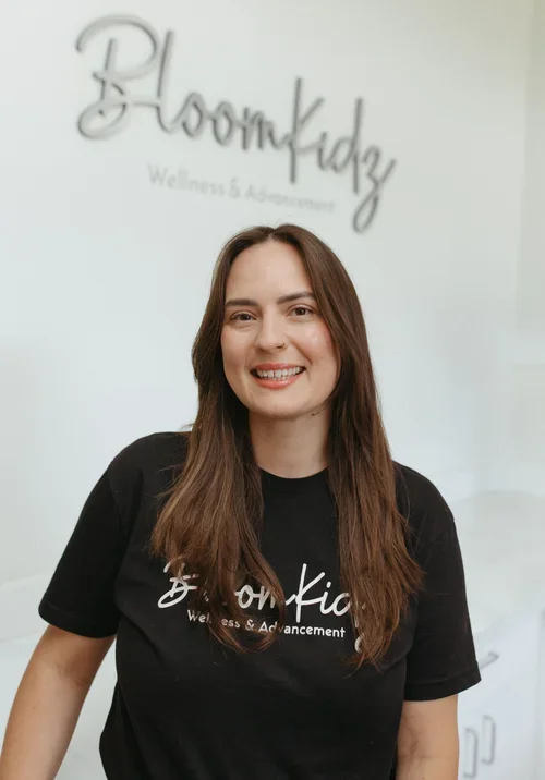 A woman with long brown hair smiling at the camera, wearing a black t-shirt with the logo 'Bloomkidz Wellness & Advancement' in front of a white wall with the same logo in the background.