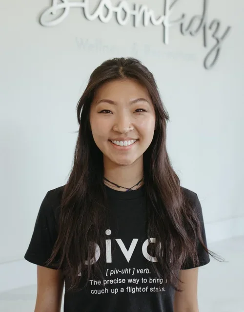 Young woman with long dark hair smiling, wearing a black t-shirt with white text, standing indoors in front of a wall with the word 'BloomKidz' visible.