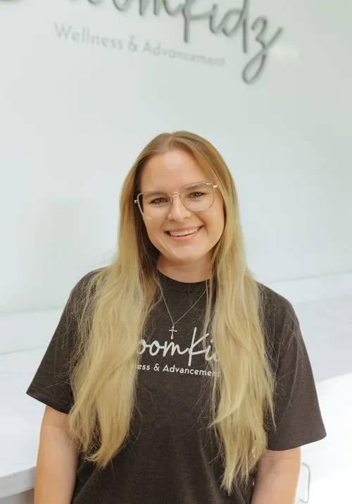 A smiling woman with long blonde hair, glasses, and a dark T-shirt standing indoors against a white wall with partially visible text in the background.