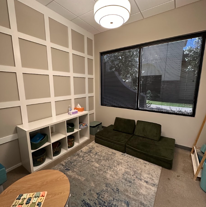 Child therapy or playroom with a dark green cushioned seating area, white storage unit with baskets, tissue box, and toys, wall with decorative molding, large window with blinds, round ceiling light, and a patterned rug.