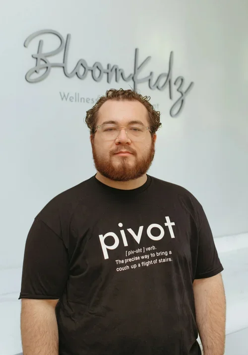 Man with glasses and a beard standing in front of a light-colored wall with a sign that says Bloomkidz Wellness & advancement. He is wearing a black t-shirt that says pivot with the definition underneath.