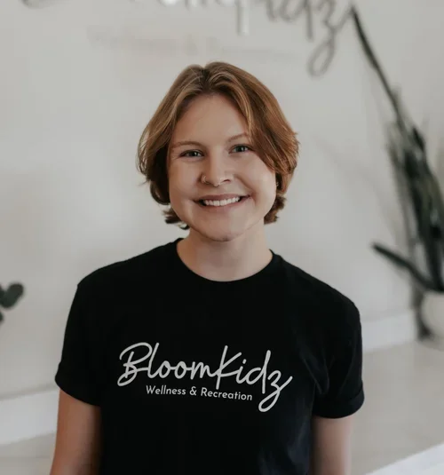 A young woman with short, light brown hair smiling, wearing a black t-shirt that says 'Bloom Kidz Wellness & Recreation'.