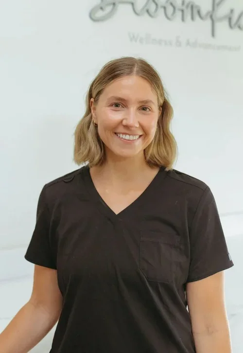 A woman with shoulder-length blonde hair, wearing a black scrub top, smiling against a light-colored background with a logo that reads 'BloomKidz Wellness & Aesthetics'.