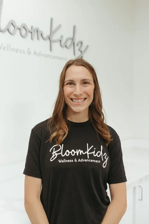 Smiling woman with brown hair wearing a black BloomKidz Wellness & Advancement t-shirt in a bright room with BloomKidz sign on the wall.
