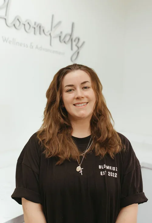 A smiling woman with shoulder-length wavy brown hair, wearing a black T-shirt with white text and a necklace, standing indoors in front of a white wall with a sign that reads 'BloomKidz Wellness & Advancement'.