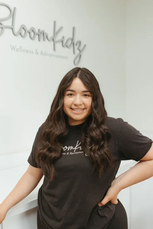 A young woman with long, wavy dark hair smiling, wearing a black T-shirt with 'BloomKidz Wellness & Advancement' printed on it, standing in a wellness center with a white wall in the background that has the same words as on her shirt.