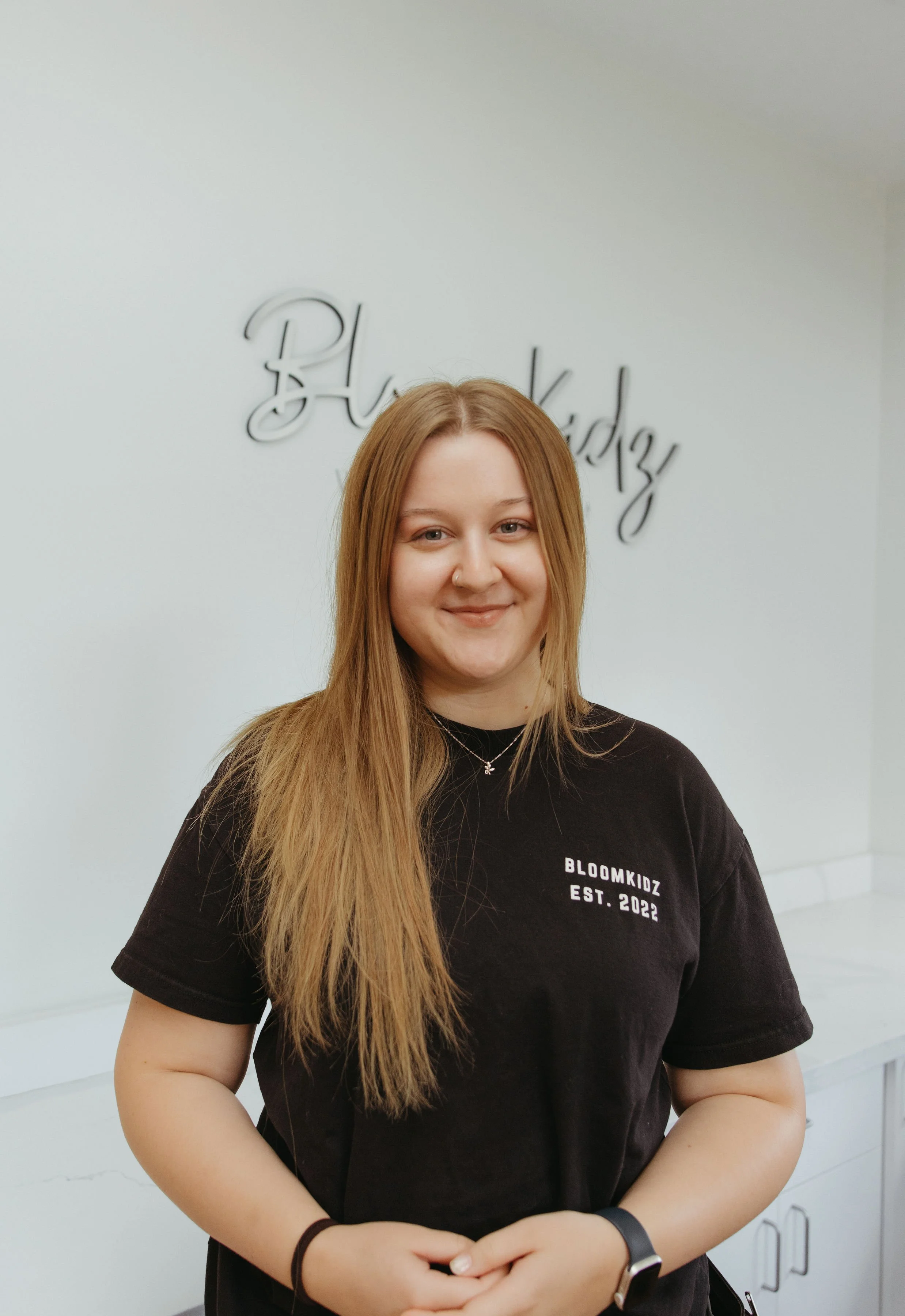 A young woman with long, light brown hair, wearing a black T-shirt that reads 'BLOOMKIDZ EST. 2022,' stands in front of a white wall. She has a small nose piercing and wears a black wristband and a watch on her left wrist.