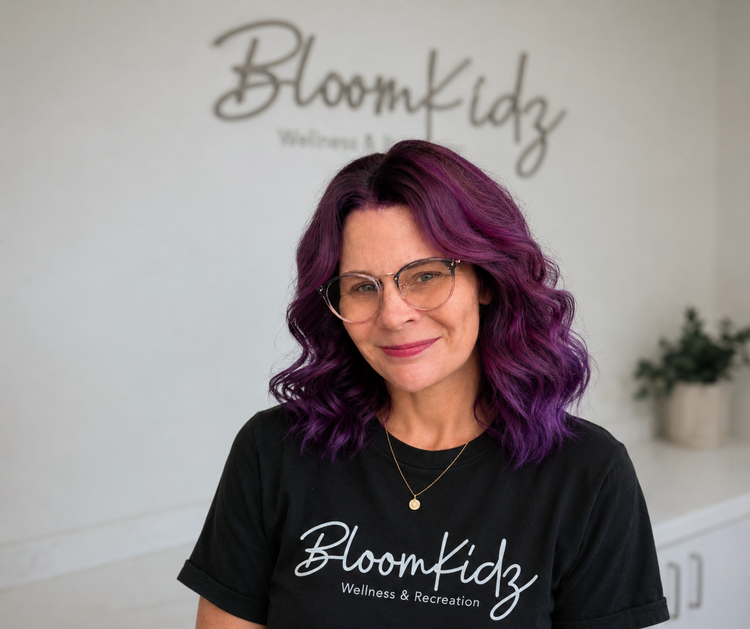Woman with purple and pink curly hair wearing glasses and a black t-shirt with the words "BloomKidz Wellness & Recreation" standing in front of a wall with the same logo.