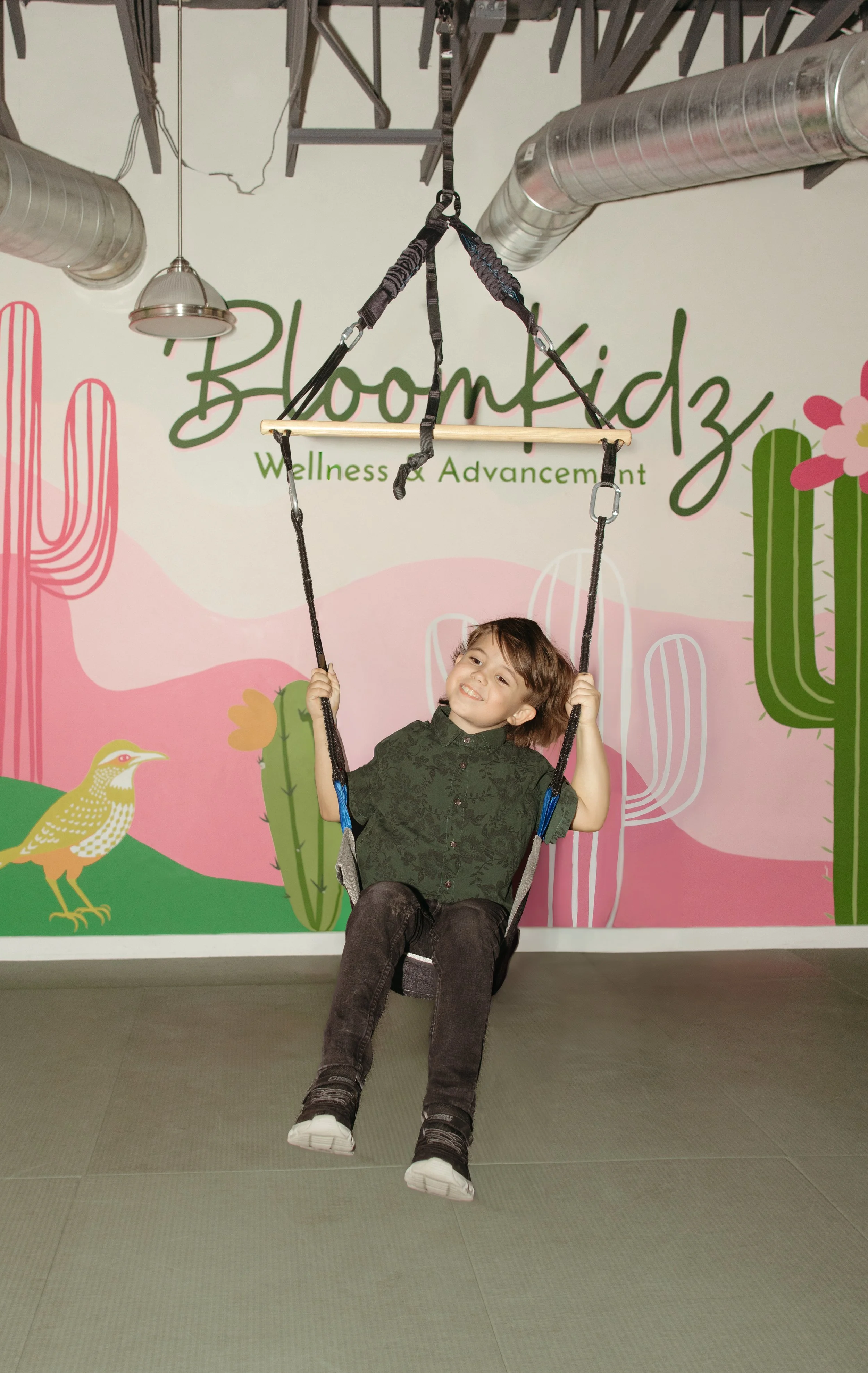 A young boy swinging on a set of indoor playground equipment. The background features colorful desert-themed illustrations and the text 'BloomKidz Wellness & Advancement'.