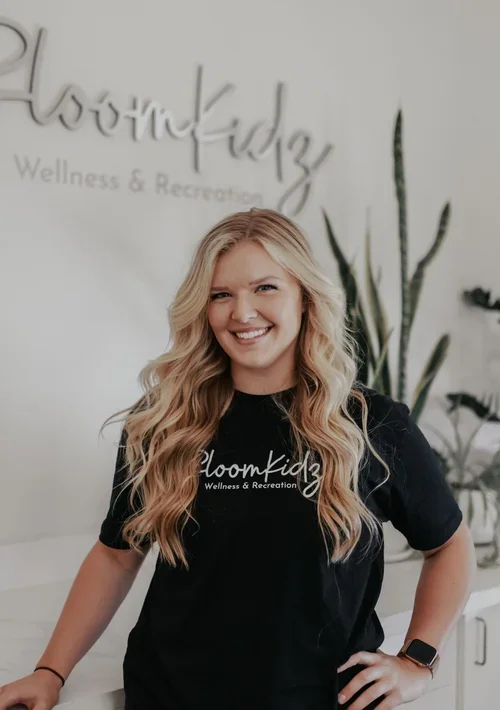 A smiling young woman with long blonde hair wearing a black Bloomkindz T-shirt, standing inside a wellness and recreation center with a white wall and green plants in the background.