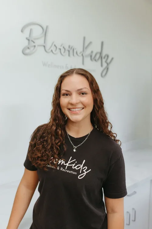 A smiling young woman with curly brown hair wearing a black t-shirt and a silver necklace, standing in front of a sign that reads 'BloomKidz Wellness & Recreation'.