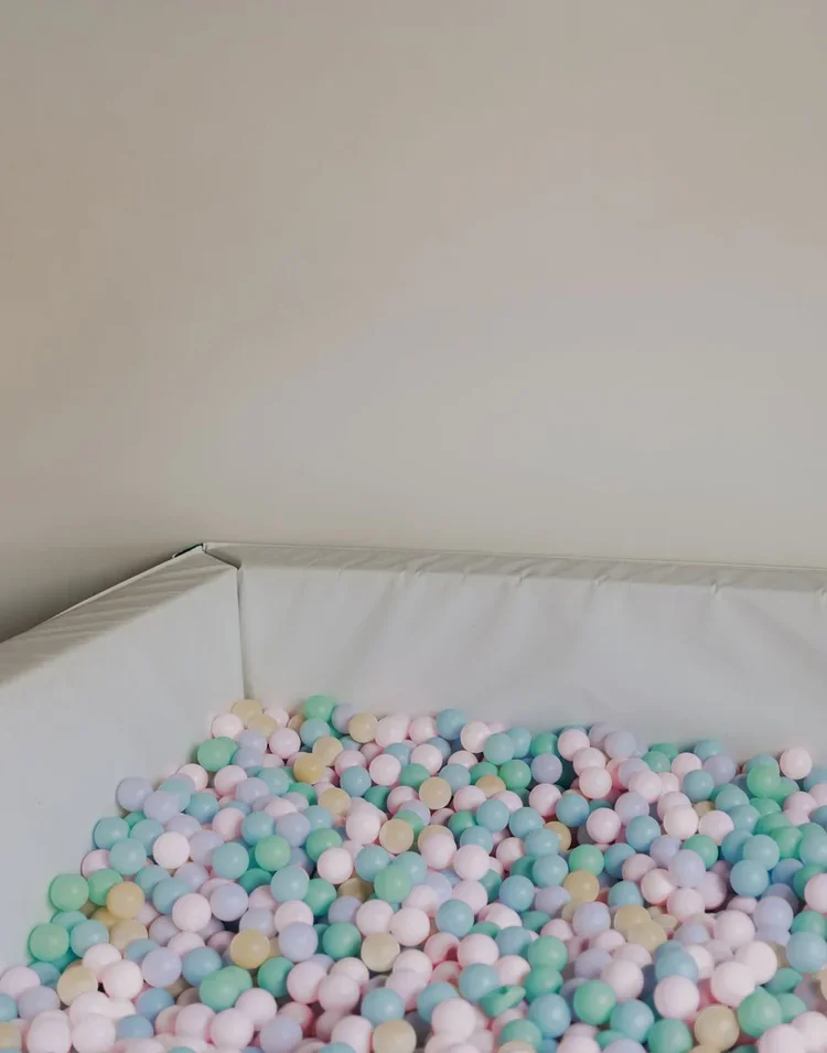 A corner of a ball pit filled with pastel-colored plastic balls in shades of pink, blue, green, and yellow, against a plain off-white wall and padded sides.