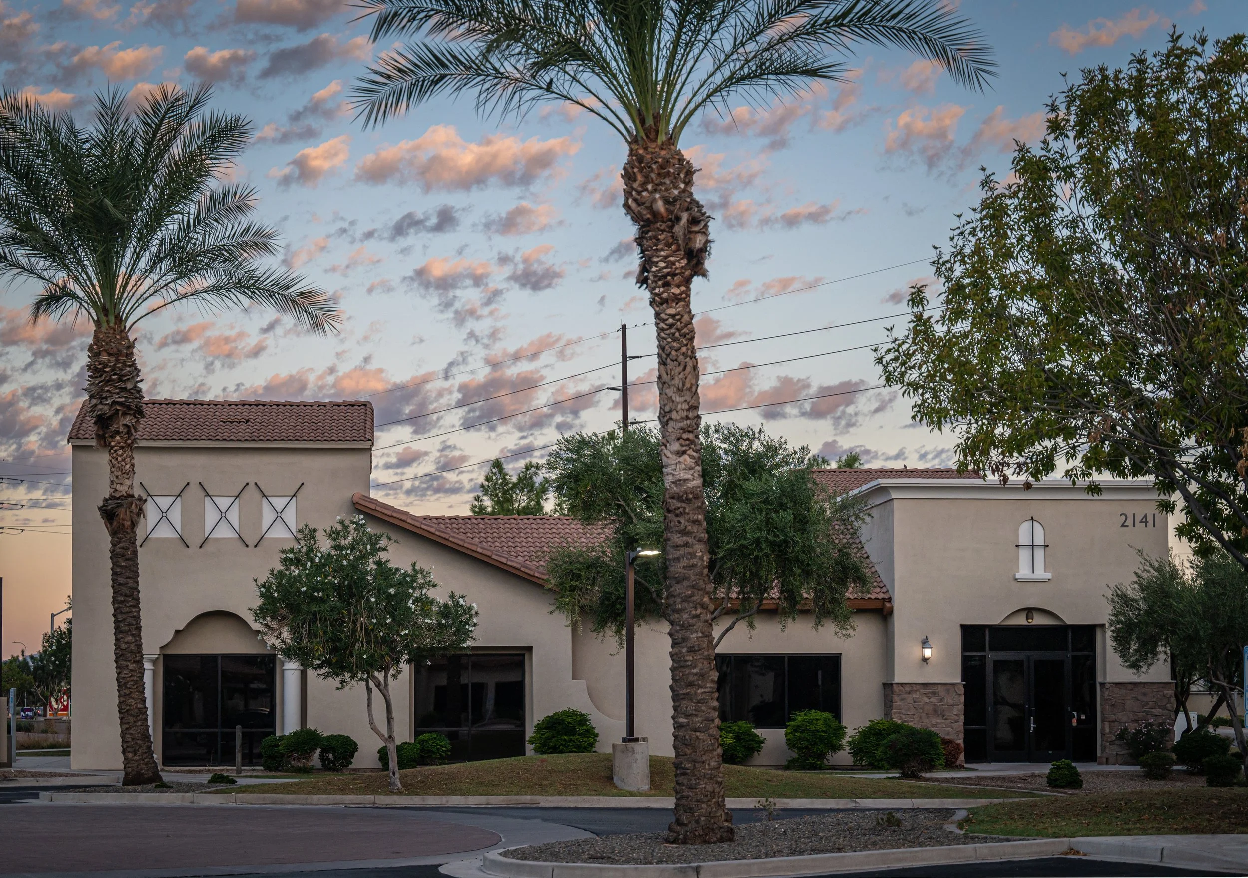 View of a medical building with white walls, red tiled roof, and windows, surrounded by palm trees and shrubbery, with a partly cloudy sky during sunset.