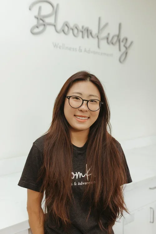 A smiling young woman with long brown hair, wearing glasses and a black t-shirt, standing in front of a white wall with the words 'BloomKidz Wellness & Advancement' written on it.