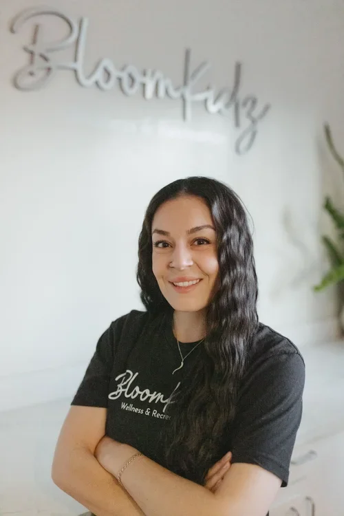 A woman with long, wavy black hair smiling and crossing her arms, standing in front of a white wall with a decorative sign that says 'BloomKidz' and a green plant on the right.