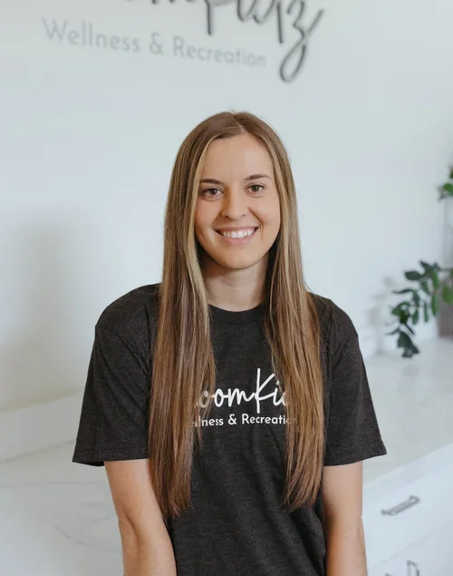 Smiling young woman with long hair wearing a black t-shirt with white text, standing in a wellness and recreation center with white walls and a plant in the background.