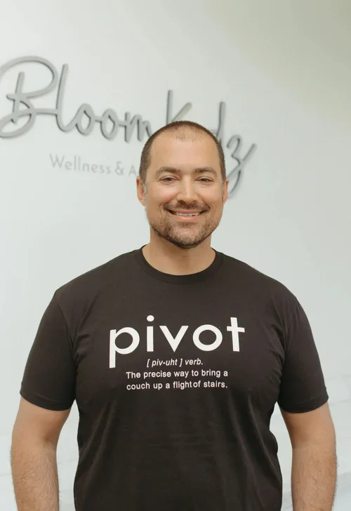 A man smiling, wearing a black t-shirt with the word "pivot" and its definition printed on it, standing in front of a white wall with partial text reading "BloomKidz" and "Wellness & Recreation".