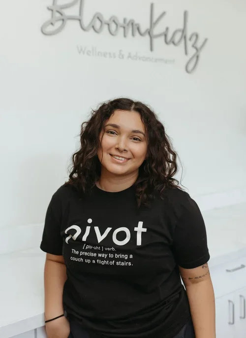 A young woman with curly brown hair smiling and wearing a black T-shirt with the word 'pivot' and a definition printed on it. She is standing indoors in front of a wall with the sign 'Bloomkidz Wellness & advancement'.