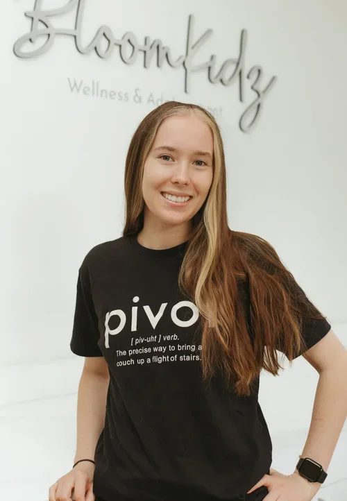 Smiling woman with long brown hair standing in front of a white wall with 'BloomKidz' written on it, wearing a black t-shirt with a definition of the word 'pivot' and a smartwatch on her left wrist.