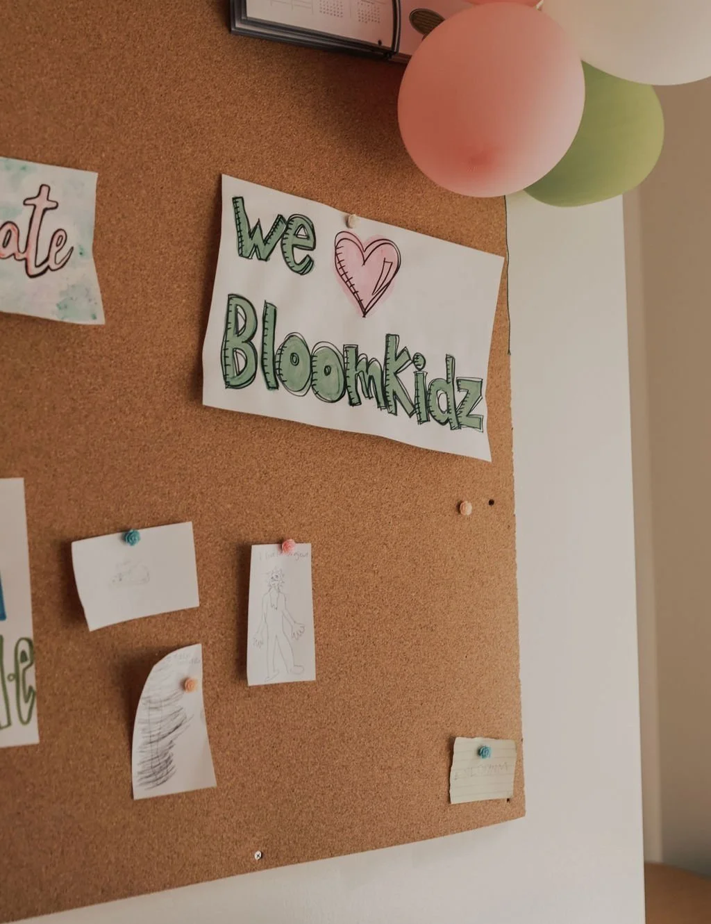 Decorated corkboard with a sign that reads 'we ♥ Bloomkidz', pink, green, and white balloons above, and various smaller pieces of paper pinned below.