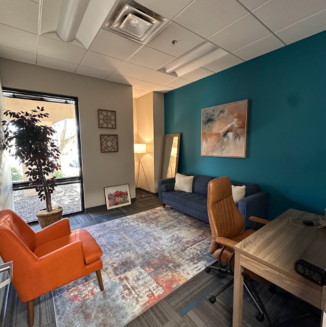 Office lounge with blue accent wall, blue sofa, orange armchair, wooden desk, standing mirror, framed artwork, floor lamp, potted plant, and window with curtains.