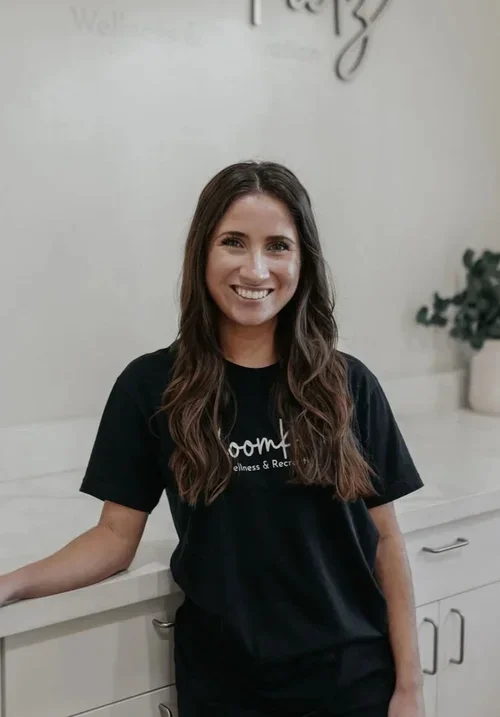 Smiling woman with long brown hair in a black shirt standing in a modern room with a white counter and a plant in a white pot.