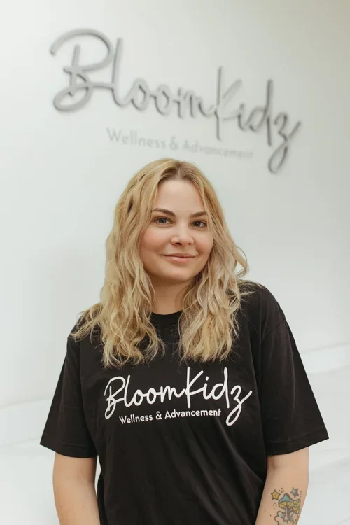 A woman with blonde wavy hair smiling at the camera, wearing a black t-shirt with white text that says 'Bloomkidz Wellness & Advancement'. In the background, there is a sign that says 'Bloomkidz Wellness & Advancement' on a light-colored wall.