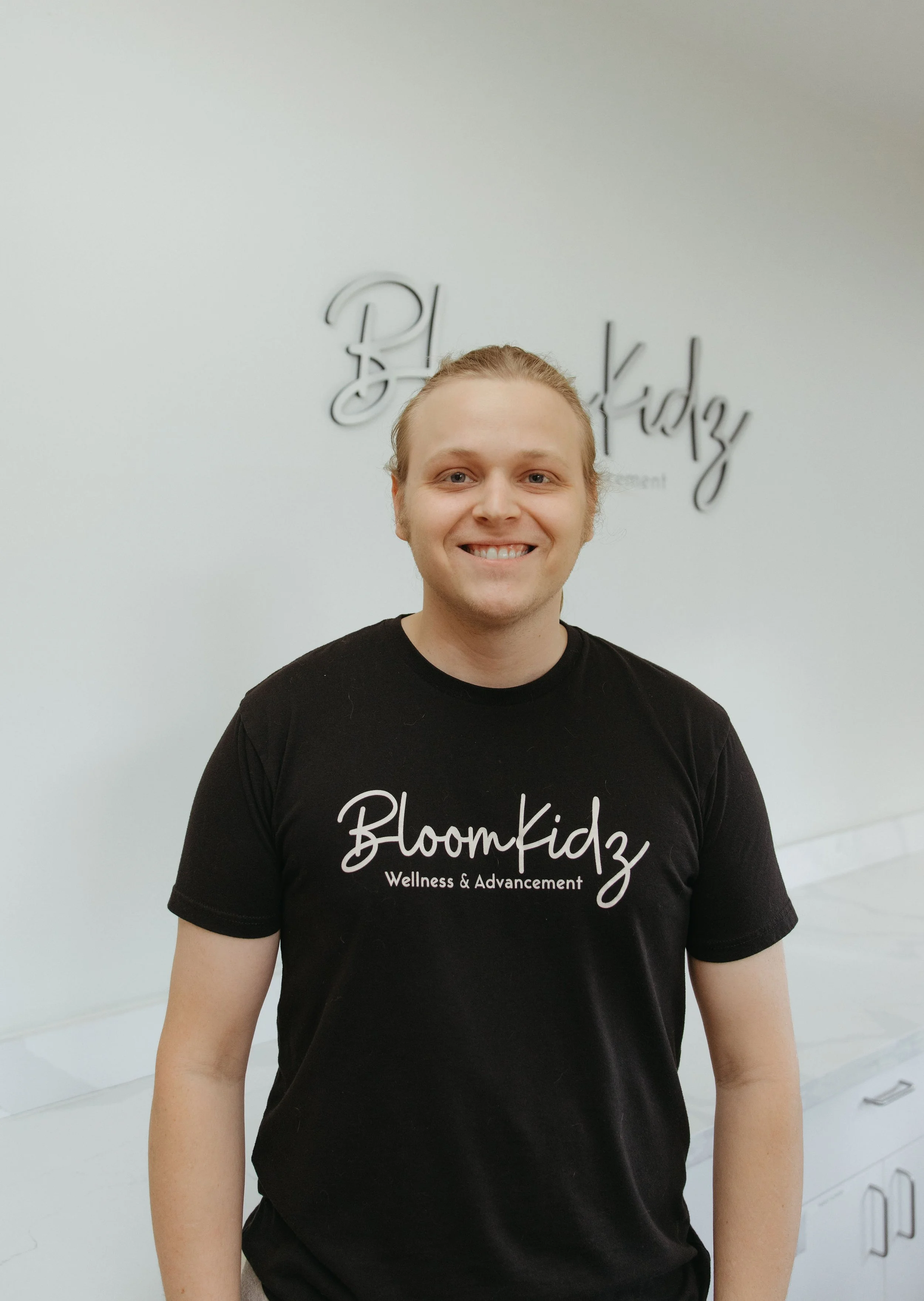 Smiling man wearing a black shirt with white text that reads "BloomKidz Wellness & Advancement" stands in front of a white wall with a decorative sign that says "BloomKidz."