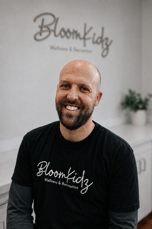A smiling man with a beard and a shaved head wearing a black t-shirt with 'BloomKidz Wellness & Recreation' printed on it, standing in a wellness center with a sign that reads 'BloomKidz Wellness & Recreation' on the wall behind him.
