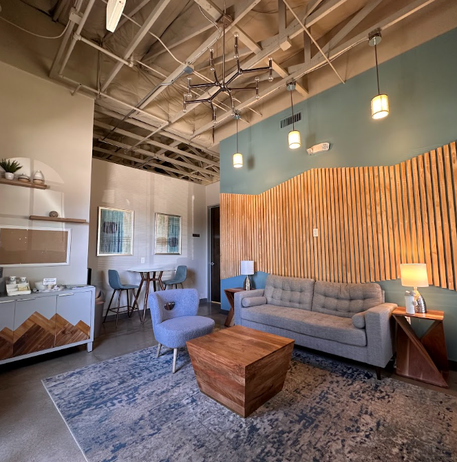 Living room with a light gray couch, a blue armchair, and a wooden coffee table on an area rug. The wall has a wavy wooden detail with a teal top. There are hanging pendant lights from an industrial-style ceiling. To the side, there is a side table w