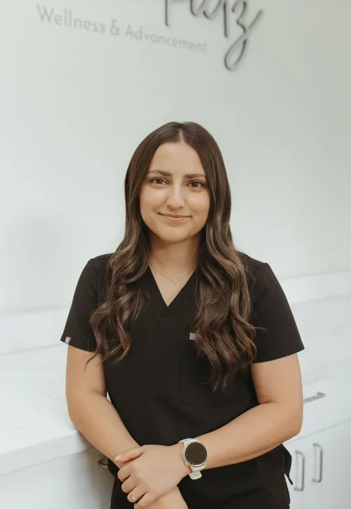 A young woman with long wavy brown hair wearing black scrubs and a smartwatch, standing in front of a white wall with 'Wellness & Advancement' in the background.
