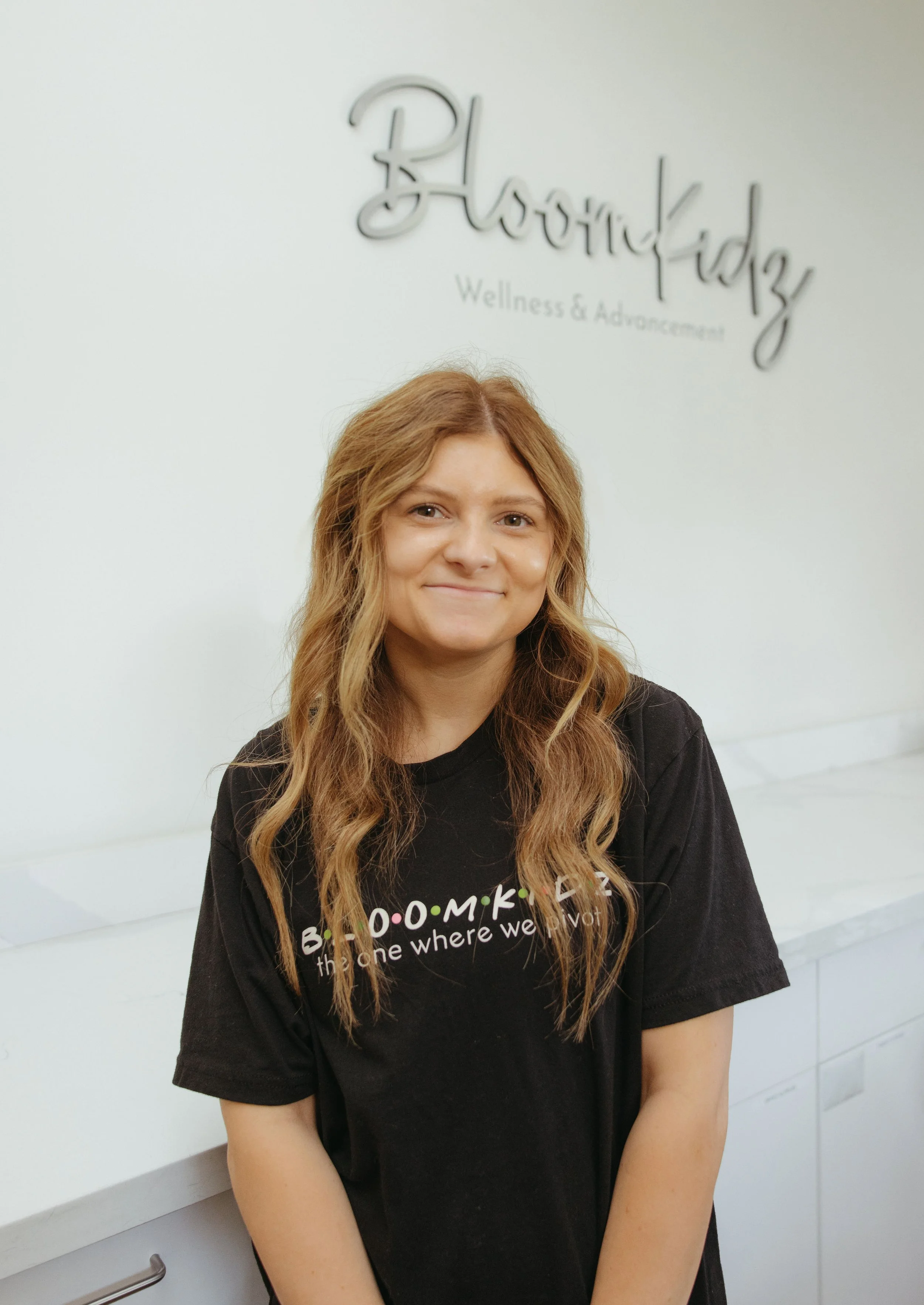 Smiling woman with long wavy hair wearing a black shirt in front of a wellness and advanced aesthetics logo