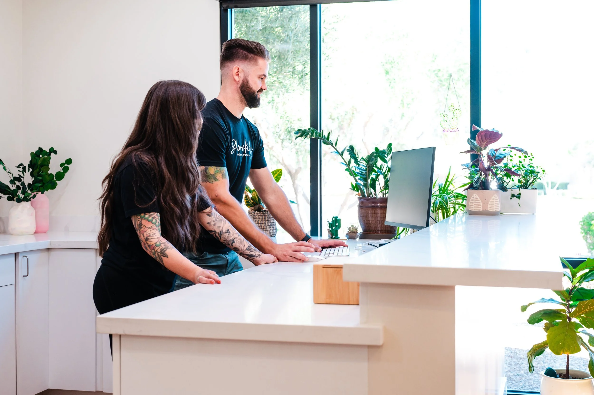 A man and a woman are standing at a counter, looking at a laptop. The woman has long brown hair and tattoos on her arm. The man has a beard and is wearing a black t-shirt. There are various potted plants on the counter and window sill, and sunlight is streaming in through large windows.