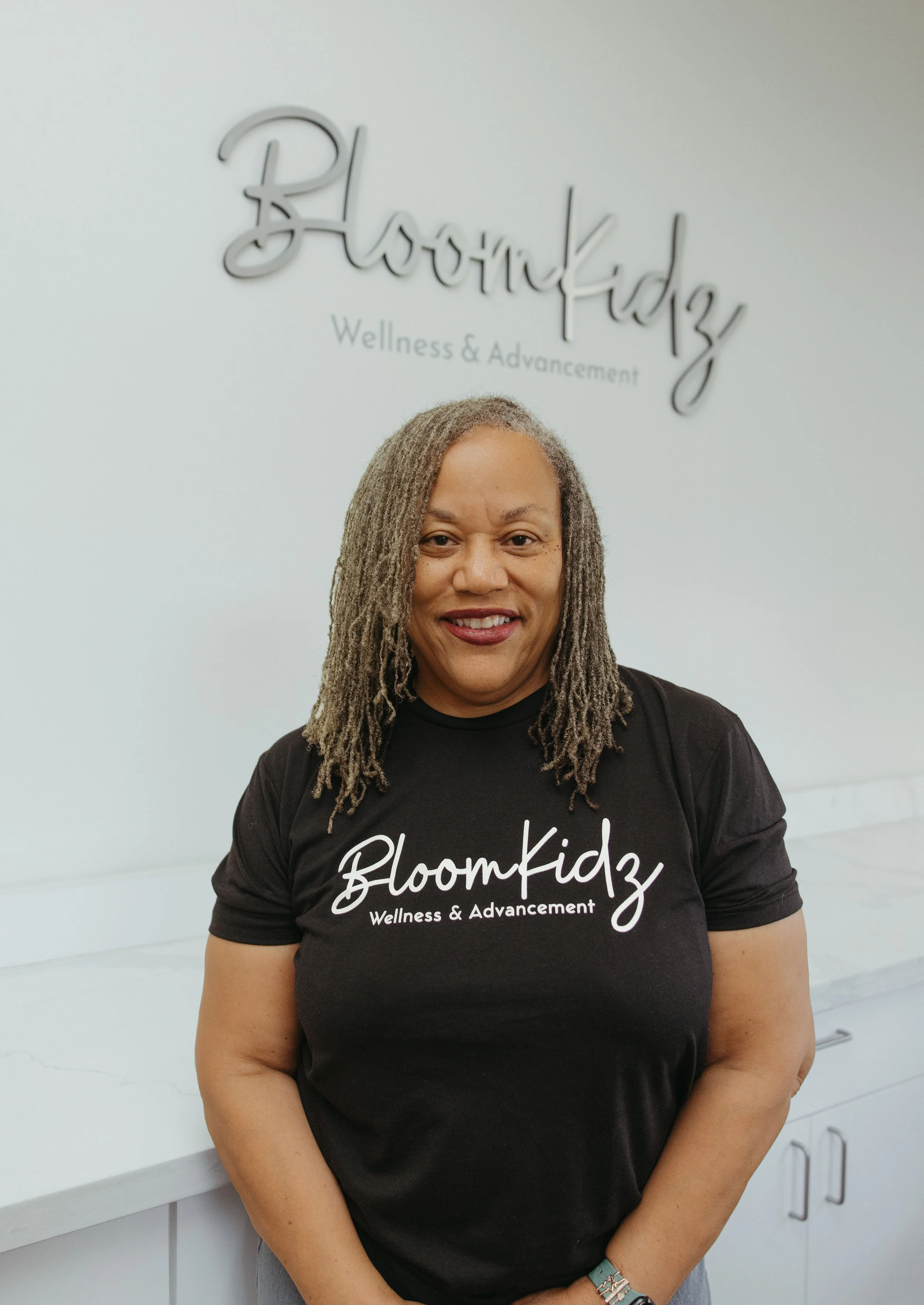 A woman with long, gray dreadlocks smiling, wearing a black t-shirt with white text that says 'BloomKidz Wellness & Advancement,' standing in front of a wall with the same text and logo for BloomKidz.