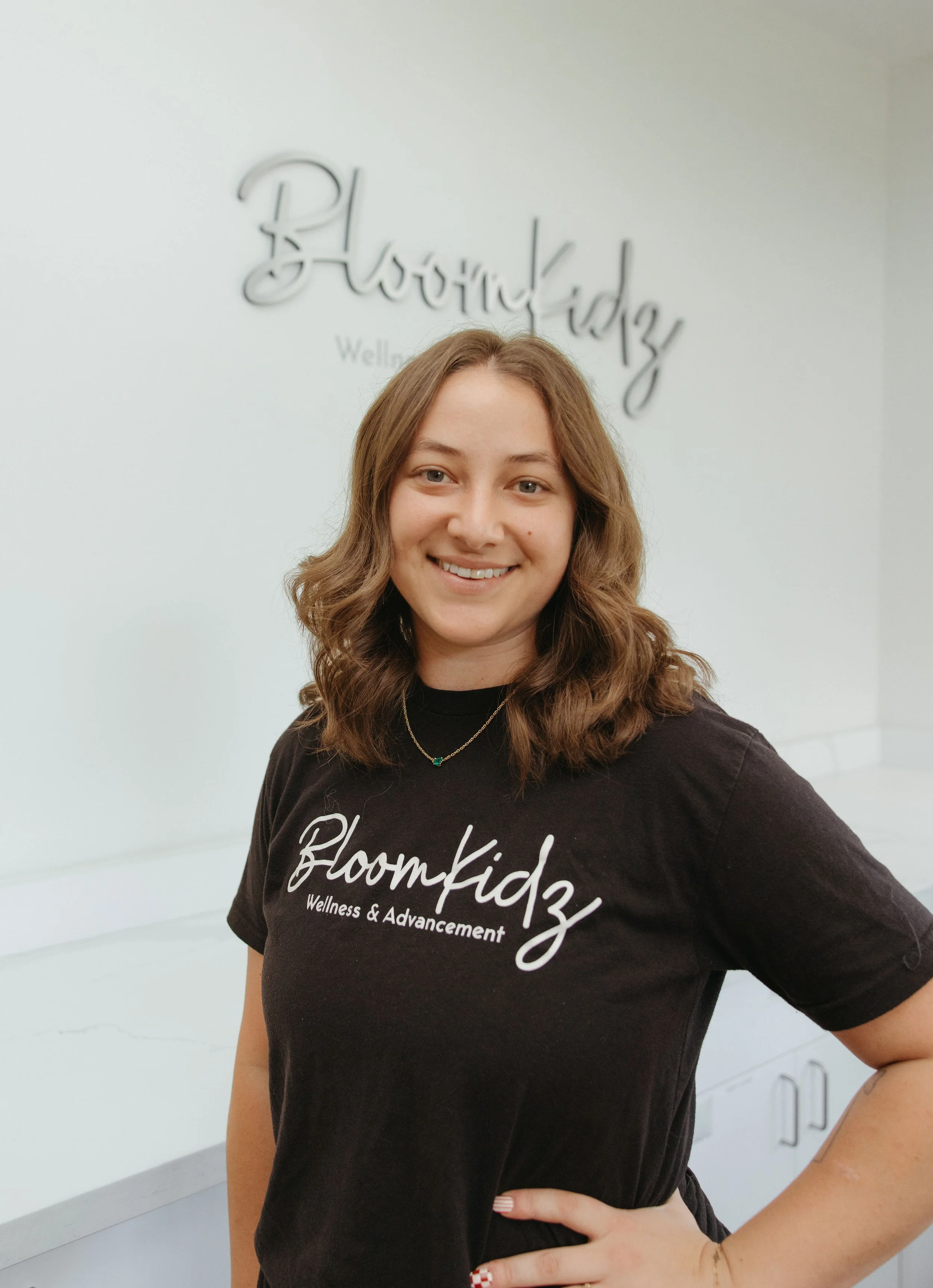 A woman with shoulder-length wavy brown hair smiling in front of a white wall with a sign that reads 'Bloomkidz Wellness & Advancement.' She wears a black t-shirt with the same text and a necklace with a small green pendant.
