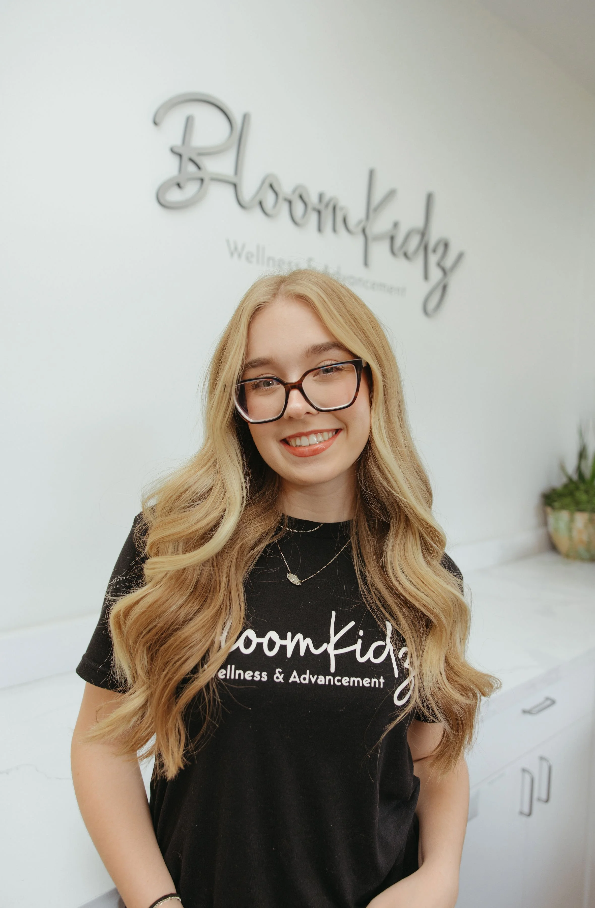 Smiling blonde woman wearing glasses and a black BloomKidz Wellness & Advancement T-shirt