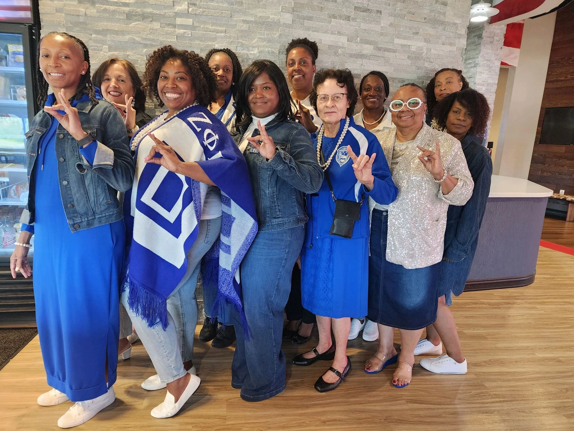 A group of predominantly African American women and a few women of different ethnicities standing indoors, smiling, posing and making hand gestures. They are dressed in blue and white, with some wearing accessories like glasses, necklaces, and scarve