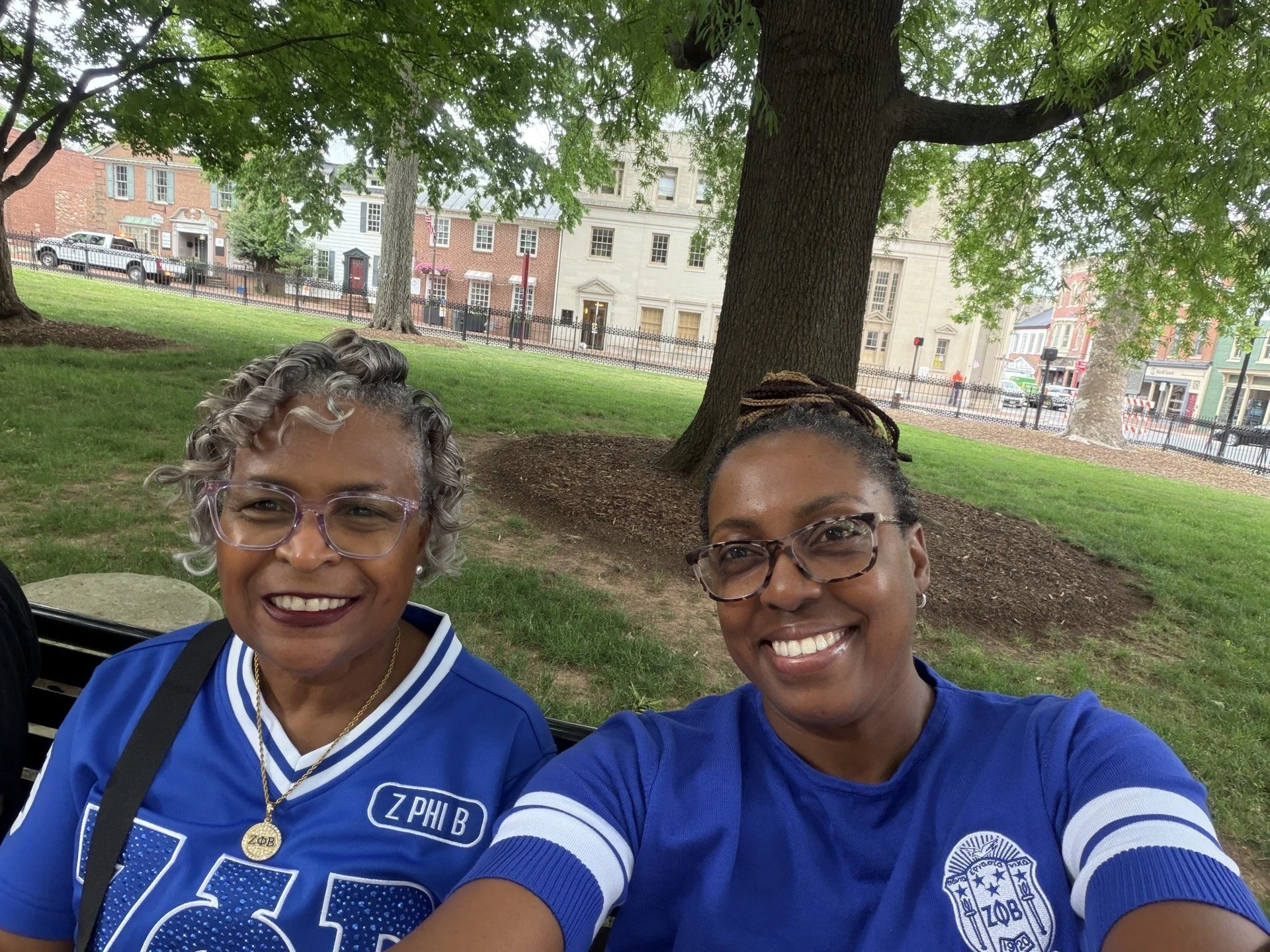 Two smiling women wearing blue sports jerseys, sitting on a park bench with a large tree behind them, in an urban park setting with colorful buildings across the street.