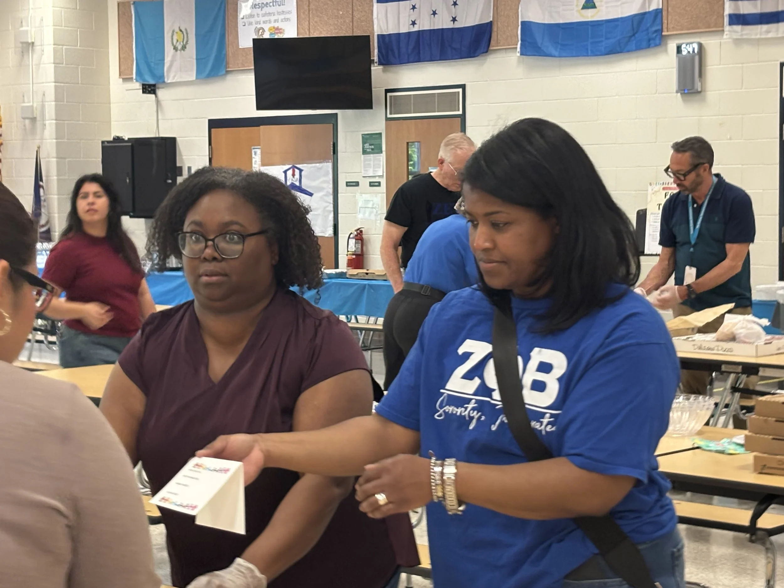 People gathered in a room decorated with flags, including the two flags hanging prominently at the top, with a woman in a maroon shirt and large glasses and a woman in a blue shirt with a name tag, engaging in an activity. There are tables with items
