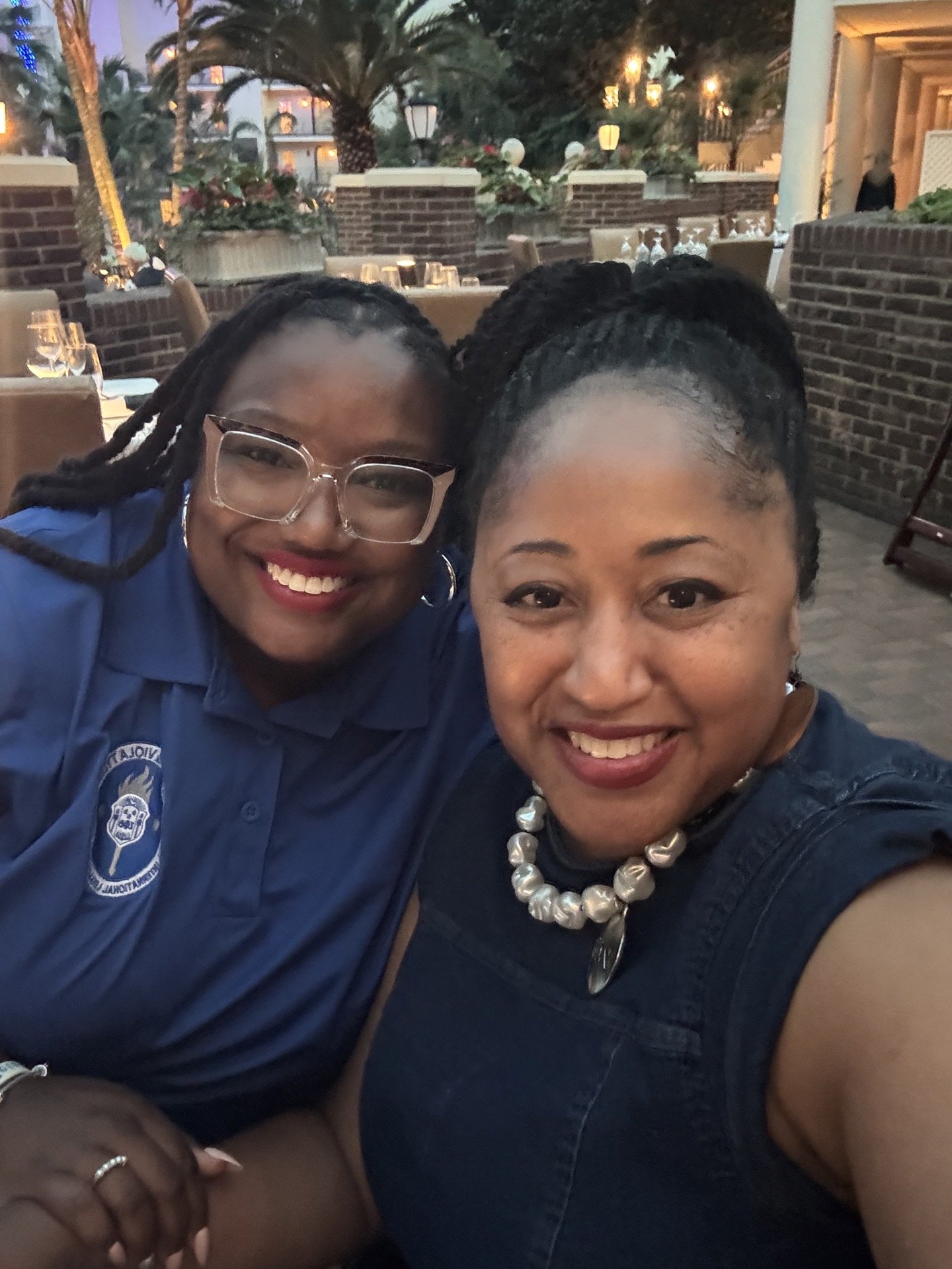 Two smiling women taking a selfie at an outdoor restaurant, with tables, chairs, and palm trees in the background, during evening time illuminated with warm lighting.