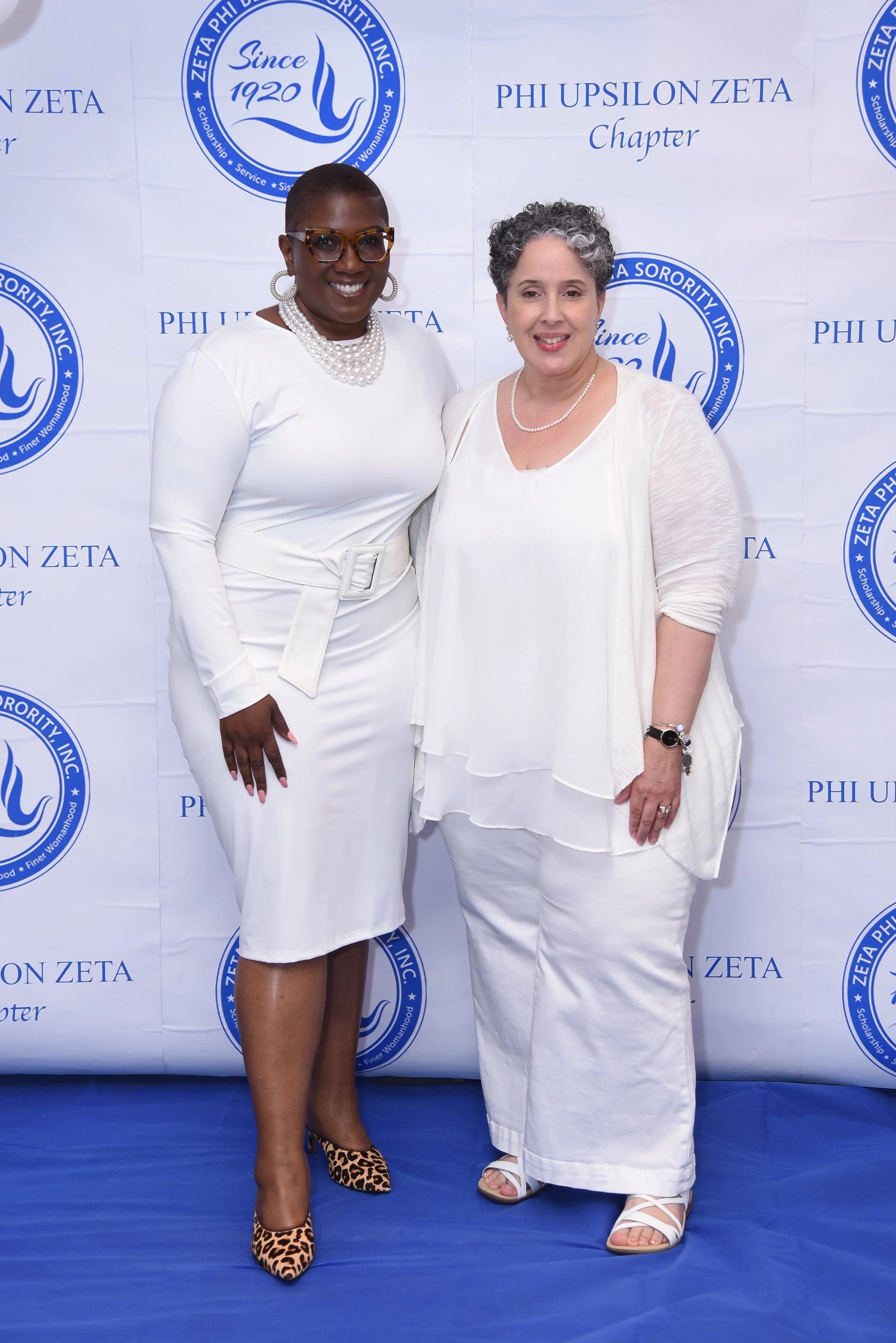 Two women standing in front of a blue and white backdrop with the Phi Upsilon Zeta logo and text. They are dressed in white outfits and smiling at the camera.