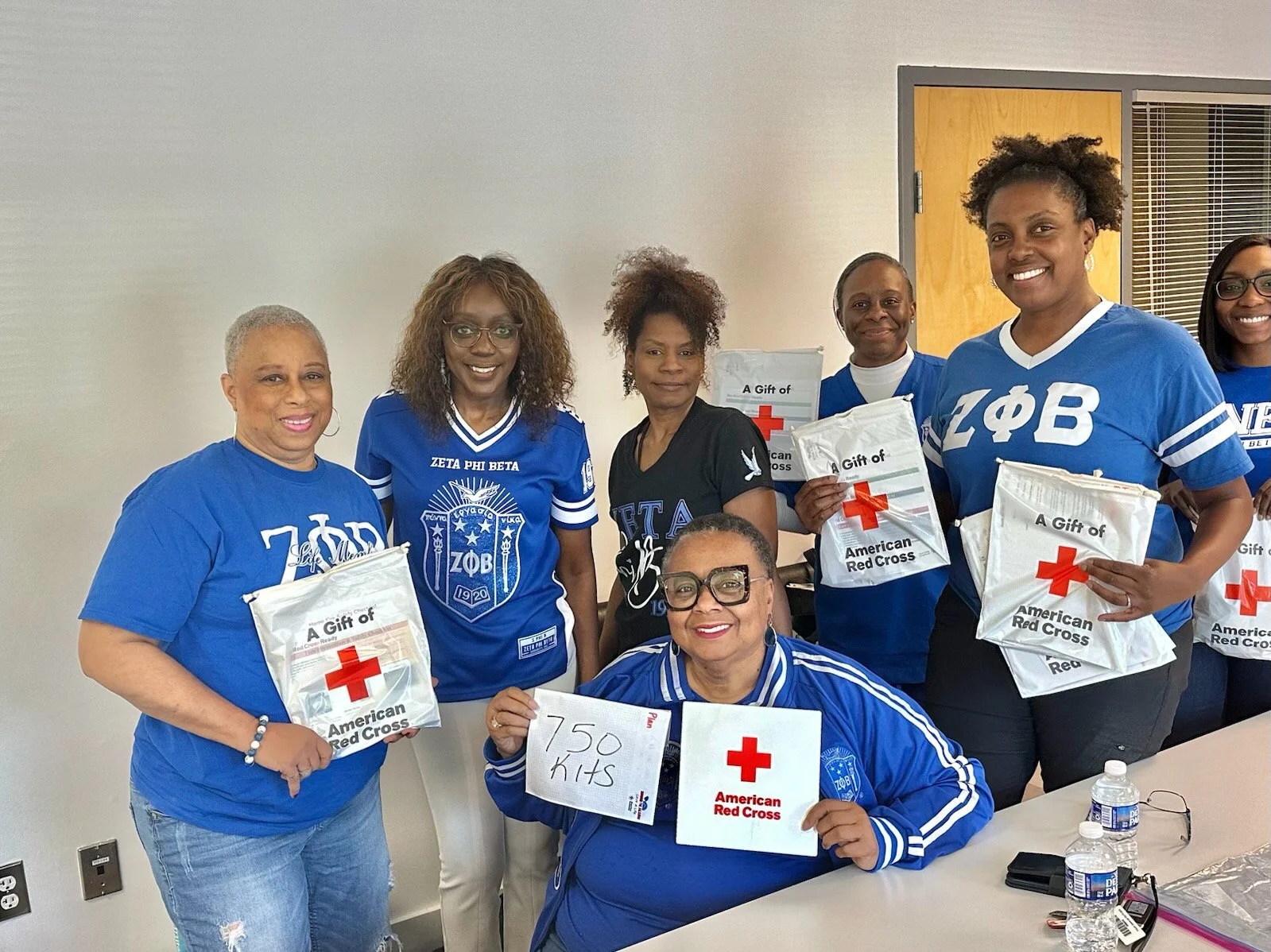 Group of smiling women wearing blue and black shirts, holding American Red Cross packages, gathered in a room with plain white walls, some sitting at a table with water bottles, and one woman in the front holding a sign that says '750 KITS'.