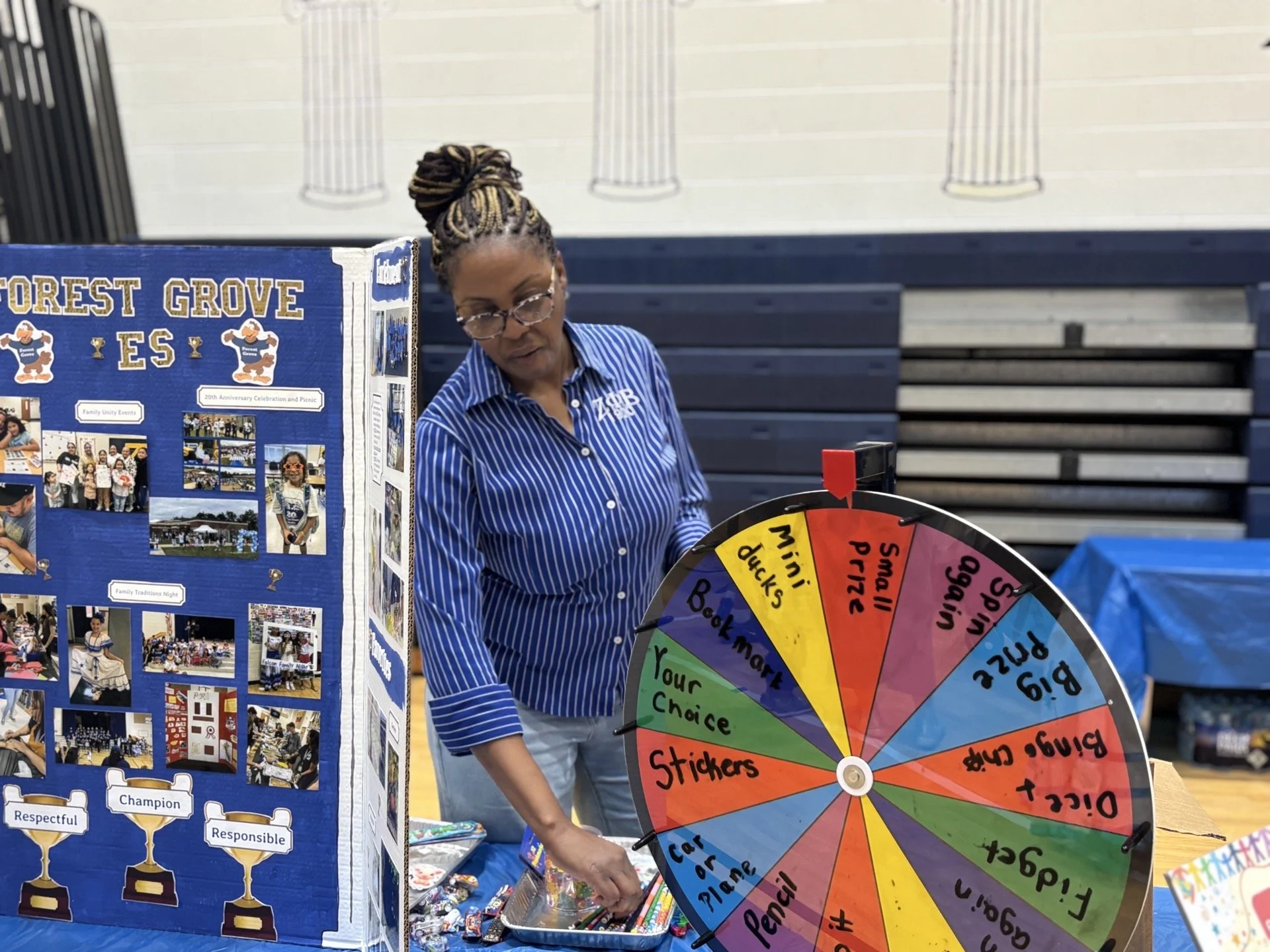 A woman wearing glasses and a blue striped shirt stands behind a colorful spinning wheel game at an event. To her left, there is a blue display board with photos and text about a celebration or event. The background includes a gymnasium with a wall a